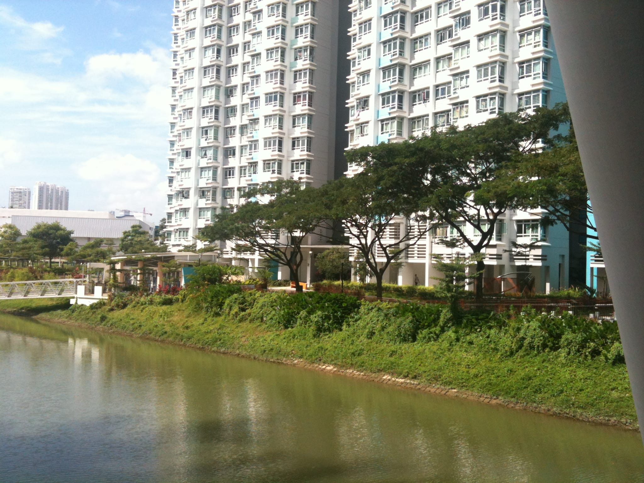 High-rise apartment buildings beside a canal lined with trees and greenery under a blue sky.